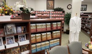 The interior of the Distribution Center underneath the Washington DC Temple. Various copies of the Book "Saints" in English and Spanish sit on a brown bookcase to the right, with a mannequin wearing a white suit standing to the right.