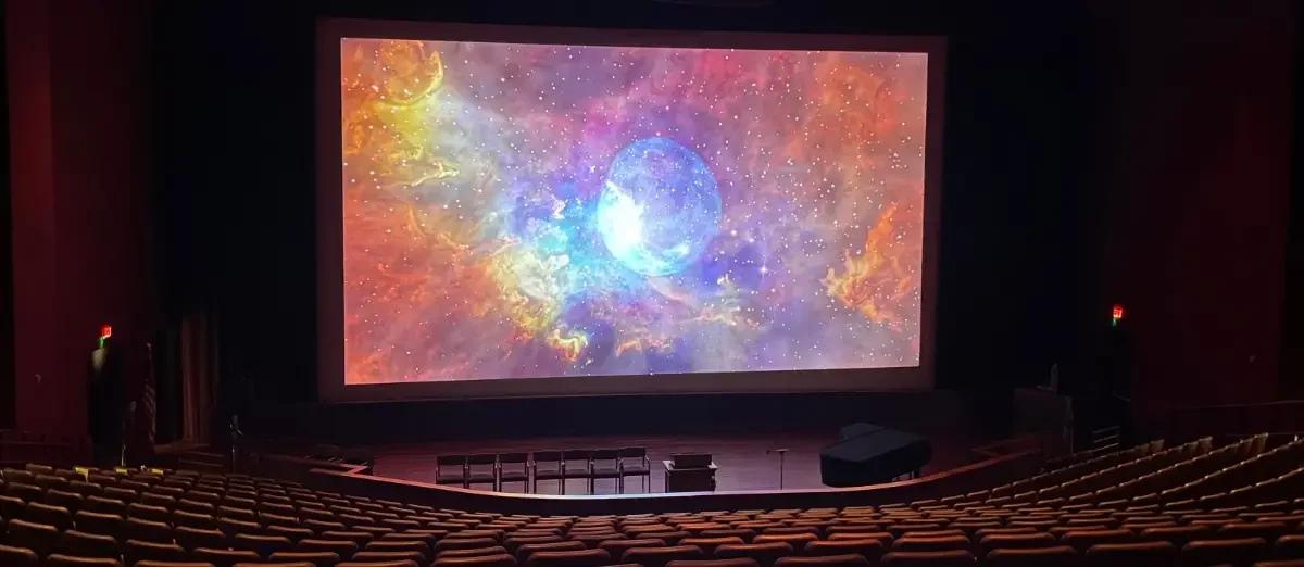 The auditorium in the Washington DC Temple Visitors' Center. The large screen displays a orange, blue, and red shot of a nebula, with a large white-blue orb in the middle of the shot.