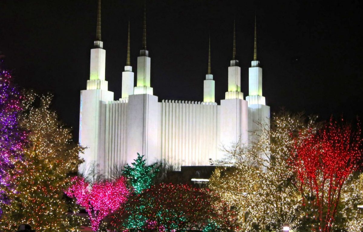 The iconic Washington DC Temple, illuminated at night during the Festival of Lights. Trees on the temple grounds are covered in Christmas lights of red, purple, pink, green, and yellow.